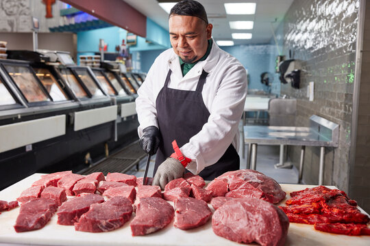 Butcher wearing hair net, apron and gloves behind counter of meat market of grocery store making cuts of steak 