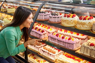 Female customer looking through display case and choosing cake from bakery counter in grocery store