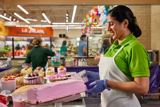 Smiling female baker spreading on strawberry frosting to decorate layer cake in latin market bakery, baker in background building birthday cake 