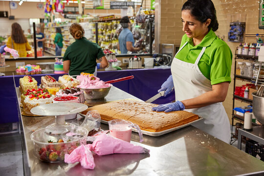 Female baker making slices of sheet cake to create layer cakes, baker in background building birthday cakes in bakery of latin super market