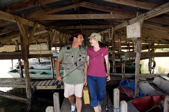 Older couple with fishing poles and accessories walking through boat dock 