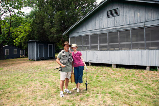 Wide angle portrait of older smiling couple in front of fishing cabin with poles and tacklebox 