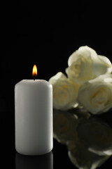 Fototapeta na wymiar White roses and burning candle on black mirror surface in darkness. Funeral symbols