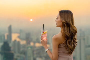 Asian woman drinking cocktail with looking cityscape at skyscraper rooftop bar in the city at...