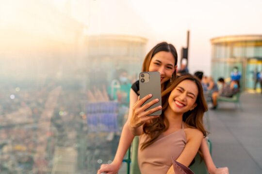 Asian woman friends enjoy urban lifestyle using mobile phone taking selfie together during have celebration dinner party at skyscraper rooftop bar in the city at sunset on summer holiday vacation.