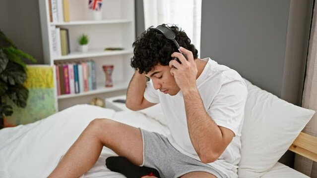Young latin man wearing headphones listening to music relaxed on bed at bedroom