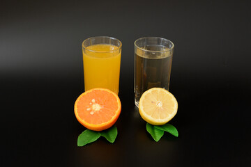 Two glasses with different citrus juices on a black background, next to pieces of ripe orange and lemon with leaves.