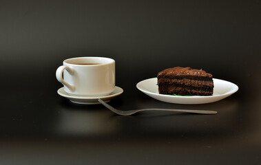 A cup of hot black coffee on a saucer, a fork and a plate with a piece of layered chocolate cake on a black background.
