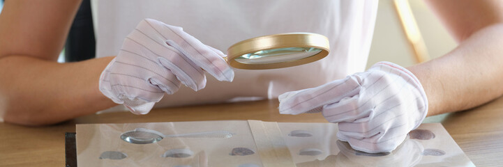 Numismatist in gloves examines collection of coins through magnifying glass.