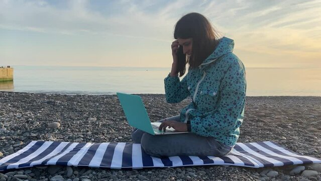 A female freelancer types a message on a portable laptop and laughs. Digital nomad. A woman during the summer holidays sits on the seashore and communicates with friends. High quality 4k footage