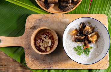 Rice porridge with fried dried fish in a white porcelain bowl garnished with coriander and red pepper on a wooden chopping board lined with banana leaves. There is a working area on the left side.