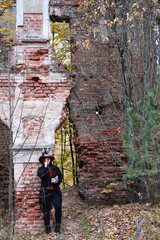 Obraz na płótnie Canvas A woman in the image of Baron Saturday poses against the backdrop of an old abandoned building with a cigar in her hands. The model is dressed in a corset, a cardigan and a top hat