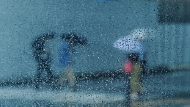 View through a glass window with raindrops on a blurred silhouette of people with umbrella walking on a rainy day. Slow motion.
