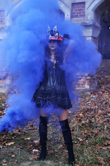 Naklejka premium A woman in a black dress with a corset and a top hat decorated with skeleton figures poses for the camera in blue smoke against the backdrop of an old abandoned building
