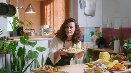 Mature female food blogger in apron putting fresh basil leaves on top of served pasta, taking plate and enjoying the smell, commenting and posing on camera with a smile in the kitchen