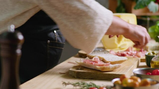 Hands of woman in apron standing by kitchen table and making gourmet sandwich with ciabatta bread, prosciutto and blue cheese. Close-up shot