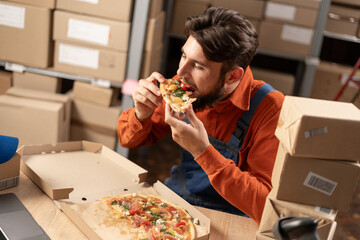 Male warehouse worker eating delicious pizza at lunch or dinner while sitting at workplace