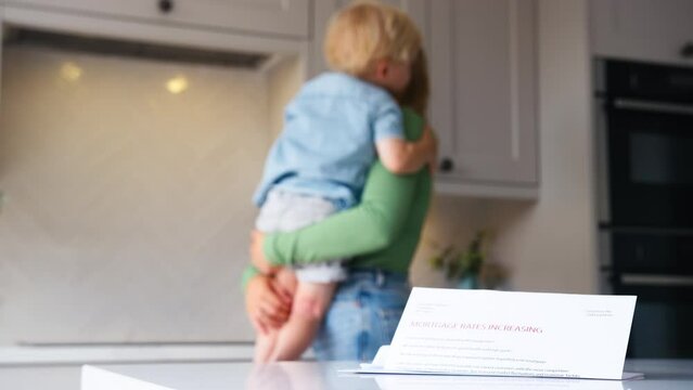 Mother cuddling young son in kitchen at home with letter about increase in mortgage rates on counter in foreground - shot in slow motion