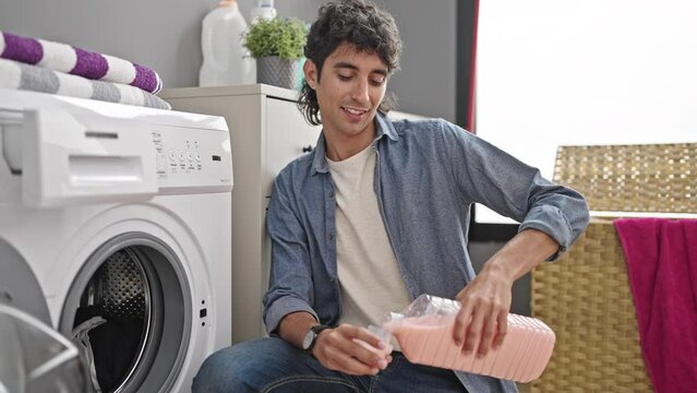 Young hispanic man washing clothes pouring detergent smiling at laundry room