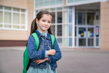 Little asian girl stands outside the school with a backpack on her back