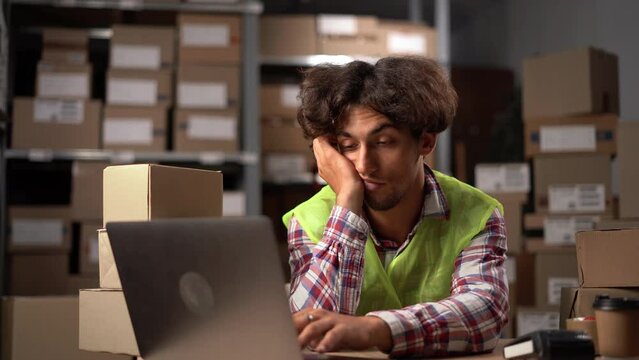 Young tired worker sitting in warehouse with laptop computer and taking a nap.