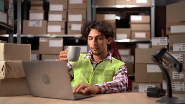 Large warehouse worker using laptop computer relaxing while enjoying hot coffee or tea