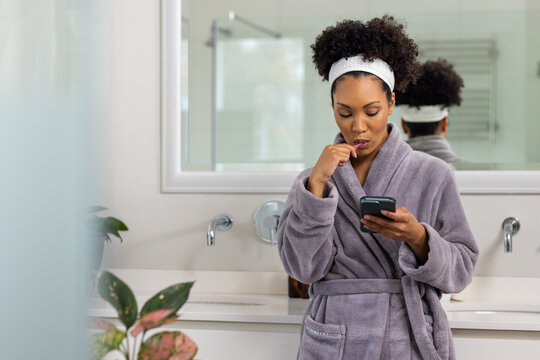Biracial woman in bathrobe brushing teeth using smartphone in bathroom, copy space - Powered by Adobe
