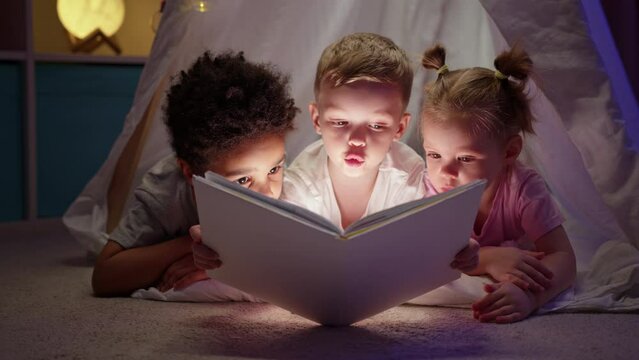 Preschooler friends at sleepover read bedtime story together in cozy play tent.