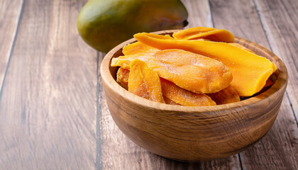 Dried mango in wooden bowl and fresh mango fruit on wood textured background. Copy space. Superfood, vegan, vegetarian food concept. Macro of orange dried mango slices, selective focus. Healthy snack.