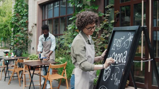 Medium shot of Caucasian waitress writing special offer on black board outside cafe while her male African American coworker preparing outdoor seatings at daytime