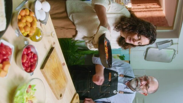 Couple of senior food bloggers taking off hot dish from oven, enjoying it, commenting on camera and adding grated cheese during online culinary class. Vertical format footage
