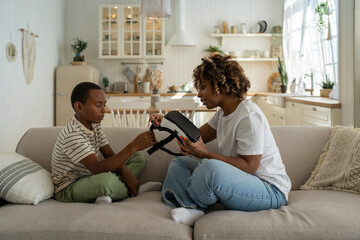 African American mother assisting son with virtual reality glasses at home, helping to use headset...