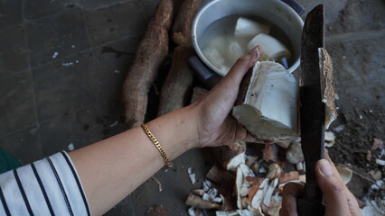 left hand with gold bracelet, holding cassava, right hand holding big knife peeling off the skin of cassava, preparation for cooking, the pile of cassava with skin that has been peeled