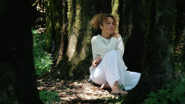 Female people in connection feeling with nature concept lifestyle. One woman sitting on the ground under a tree in the forest. Wellbeing and positive life. Mindful emotion. White clothes. Tourist