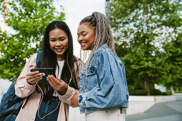 Two beautiful women watching videos with earphones and smartphone while standing at street