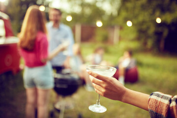 Focus on female hand holding glass of delicious cocktail. Blurred people on background. Friends meeting outdoors, picnic in nature. Concept of friendship, leisure time, weekends, summer, party