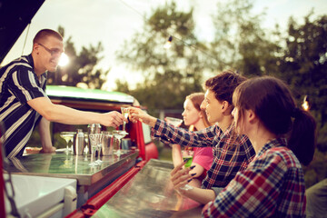 Young cheerful people, friends standing in van with guy making delicious cocktails in it. Good time at outdoor party. Concept of friendship, leisure time, weekends, summer, party