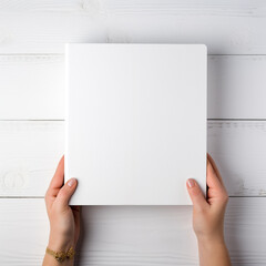 Women's hands hold a white card (box, book)  on a white wooden   background. Mockup, background for publications, presentations