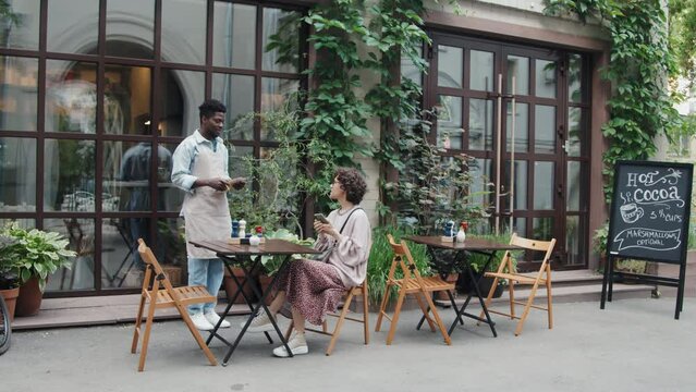 Wide shot of hospitable African American waiter coming to outside table taking customers order and making notes at daytime