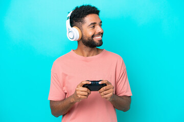 Young brazilian man playing with a video game controller isolated on blue background looking to the side and smiling