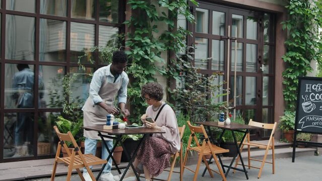Wide shot of African American waiter coming to outdoor table giving tablet with menu to Caucasian woman and writing down her order at daytime