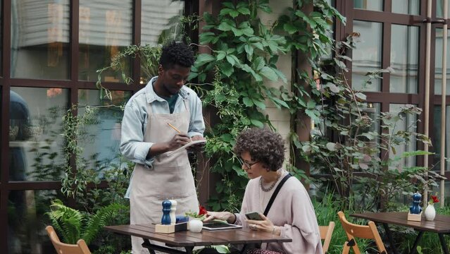 Medium full shot of Caucasian woman sitting at table on restaurant terrace choosing and ordering from menu in tablet, African American waiter taking order and writing it down