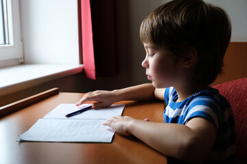 One kid sits at the table and looks at copybook and reads a task. The child is doing lesson. Correct sitting posture. There is motivation. Home education