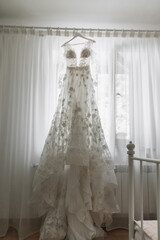 A lace dress embroidered with flowers with a train hangs on the eaves near the window in the bride's room.