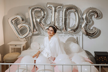 A beautiful brunette bride in a white robe, a veil and a tiara on her head, sits on a bed. Inscription bride. Happy young girl under the glow of sunlight. wedding concept