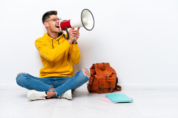 Young student caucasian man sitting one the floor isolated on white background shouting through a megaphone