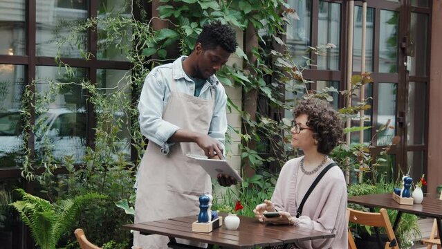 Medium shot of male African American cafe worker holding tablet with menu and helping female Caucasian customer with order on terrace outdoor at daytime