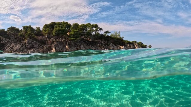 Crystal clear sea water in Sitonia, Halkidiki, Greece. Shot from border of air and water with dome. Half underwater slow-motion view of sandy beach bottom and rock with trees