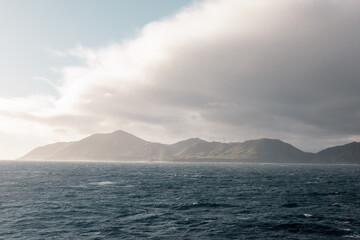 A rugged and moody looking coastline.  The coast is in Wellington New Zealand. On the hills are a number of windmills