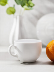 White ceramic cup and orange on the background of blurred kitchen utensils on the table, template for the designer